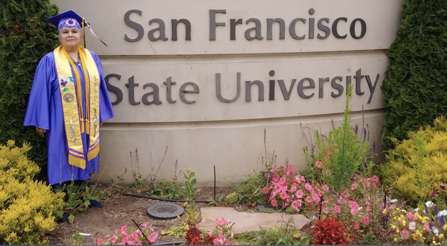 Cathi Manuel wearing cap and gown in front of SFSU monument sign