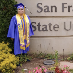 Cathi Manuel wearing cap and gown in front of SFSU monument sign